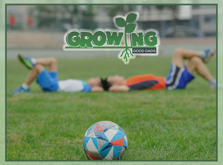 Behind a soccer ball, a dad and his son lay in a green field to look up at the sky.