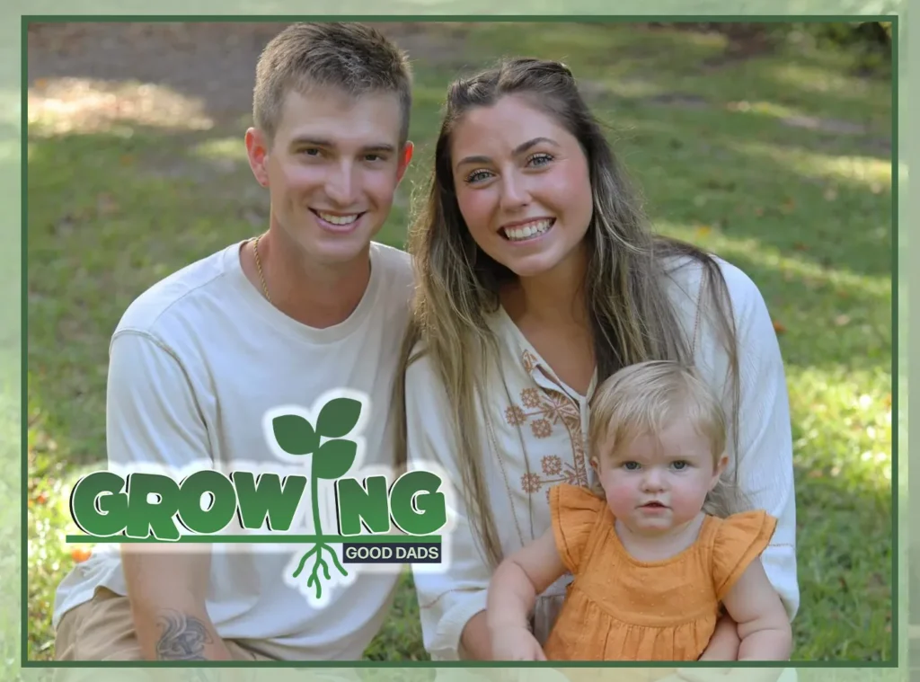 Dad Cody Dodd smiles in a family photo with his wife and young daughter while sitting on the grass together.