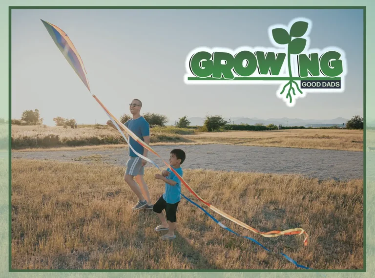 A happy man and a young boy stand in a large, empty field and hold a colorful kite.