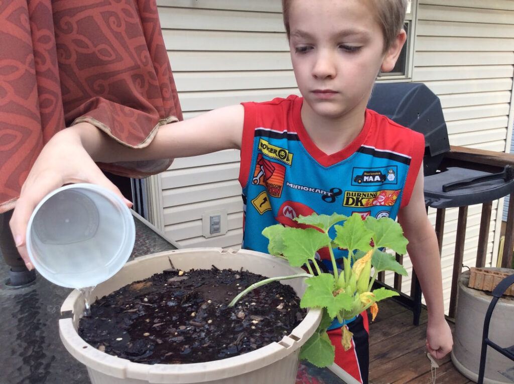 A young boy holding a plastic cup on his patio to carefully water a potted plant.