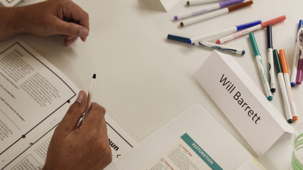 A top-down view of Will Barrett's hands on a table, holding a pen next to some markers and his name tag.