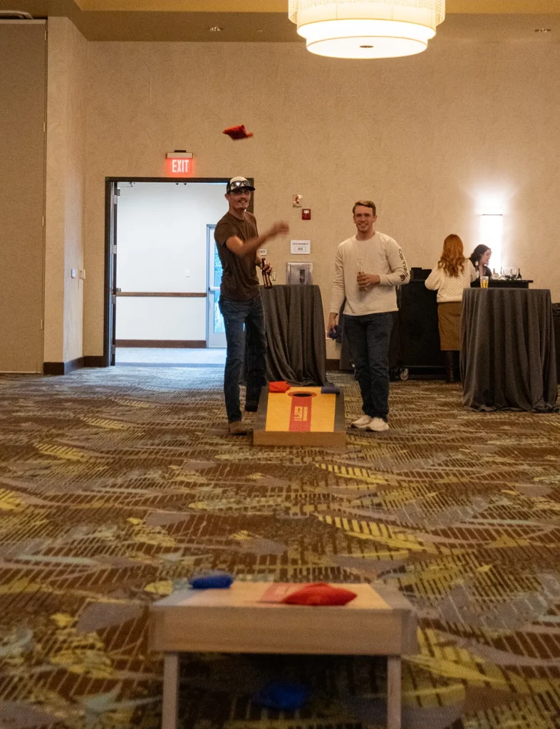 Two happy men holding beers toss a bag in a game of cornhole.