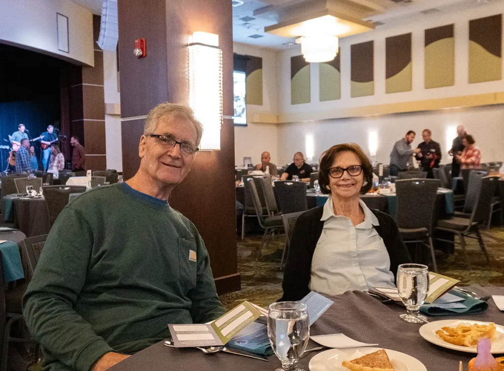 Two happy older people smile while sitting at a table while the band plays music behind them.