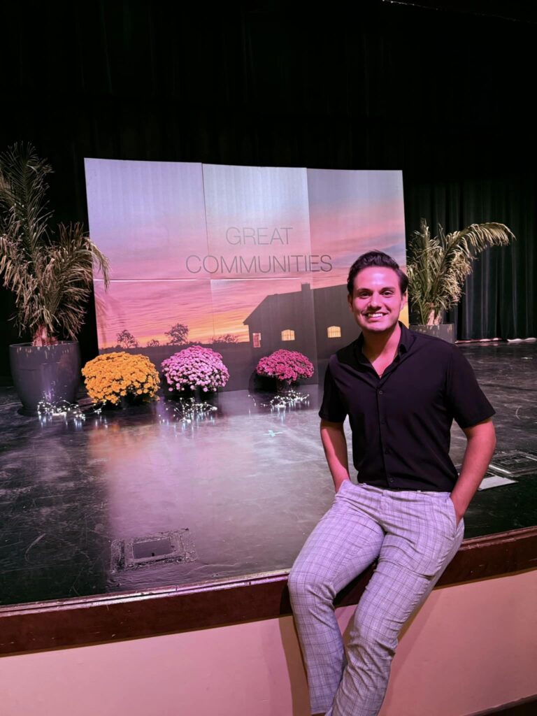 A young man sits on the stage in front of a Great Communities backdrop