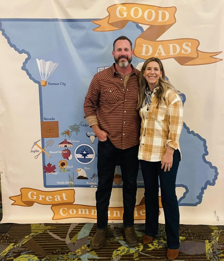 A husband and white stand in front of a banner and smile.