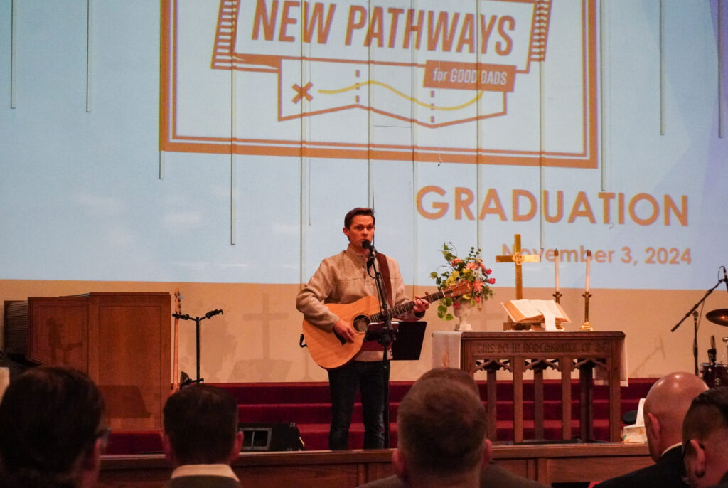 Brian Mattson stands at the front of the church's sanctuary, holding an acoustic guitar and singing into a microphone.