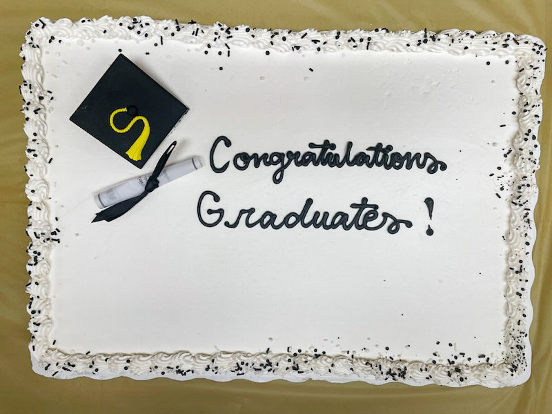 A top-down image of a rectangle cake with white frosting and black cursive writing that says "Congratulations Graduates" and a prop graduation cap and diploma.