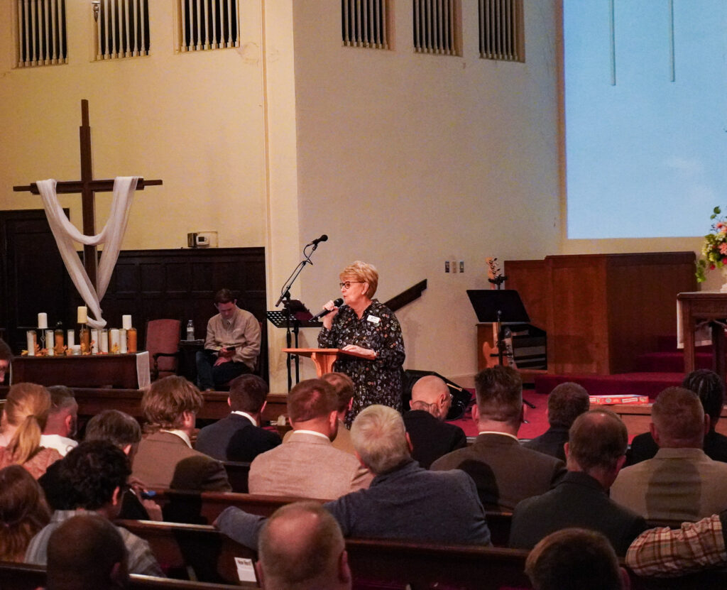 Dr. Jennifer Baker stands at the podium at the front of the sanctuary and speaks into a microphone in front of the audience in the church pews.