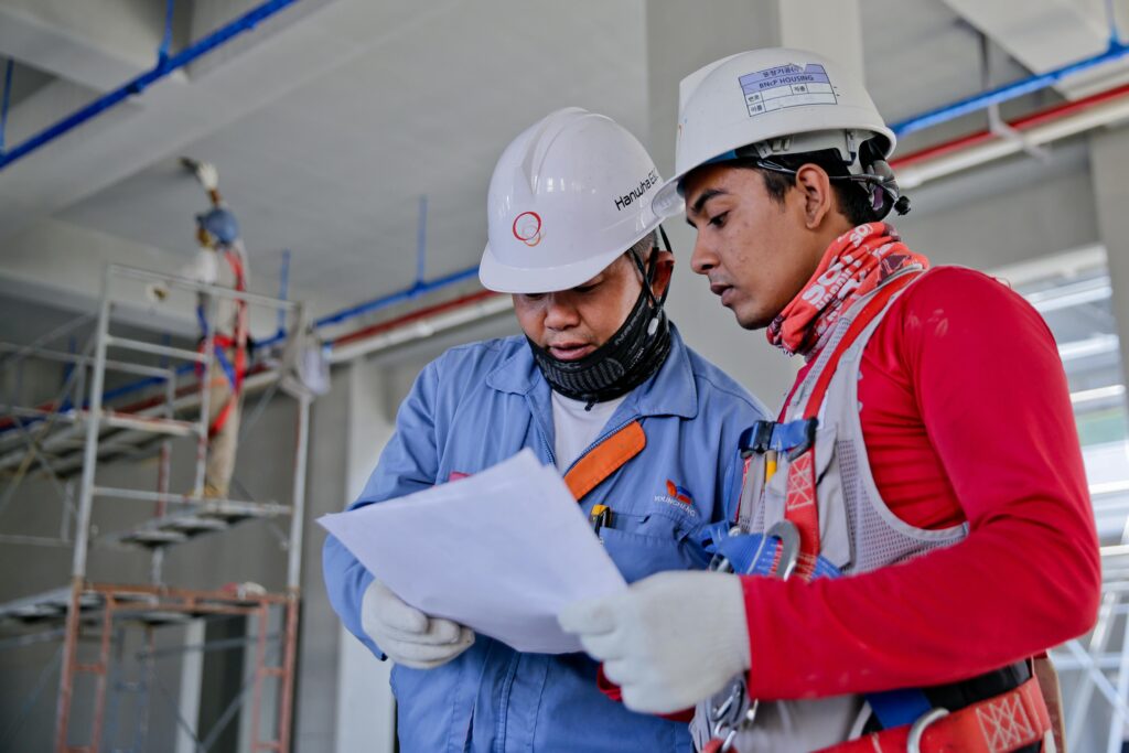 Two men in hard hats hold a piece of paper, reading it together 