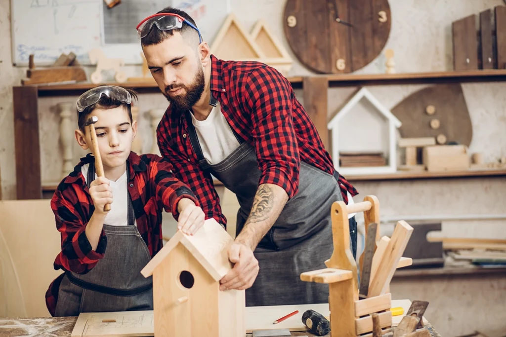 A dad and his child wear matching red plaid shirts and work on building a wooden birdhouse together as they stand in a workshop.