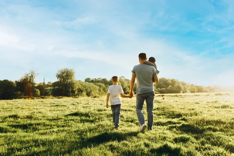 A dad and two children hold hands as they walk in a grassy field below a blue sky