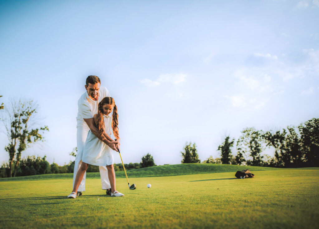 a father playing golf with his little daughter on a green field and blue sky.