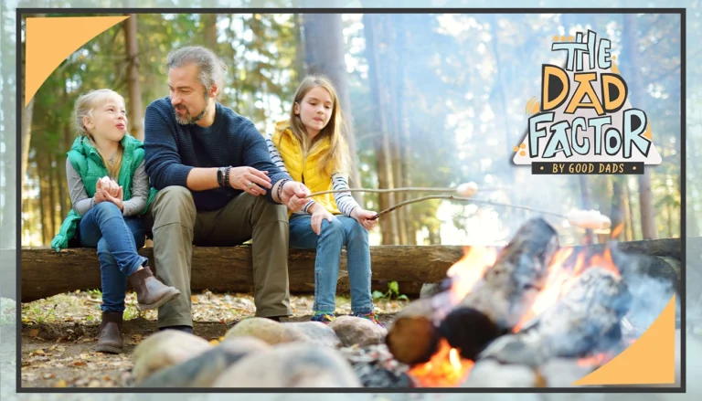 A father sits side by side with his two young daughters in front of a campfire. They are holding sticks with marshmallows.