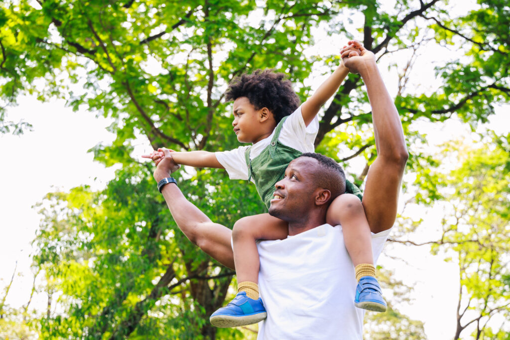 A happy Black dad holds his cute toddler son on his shoulders under a canopy of green trees