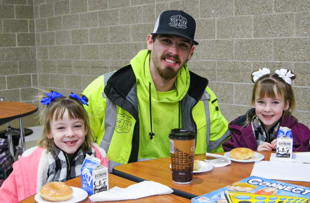 A dad and two daughters sit at a school lunch table to participate in a Good Dads Strong Schools activity
