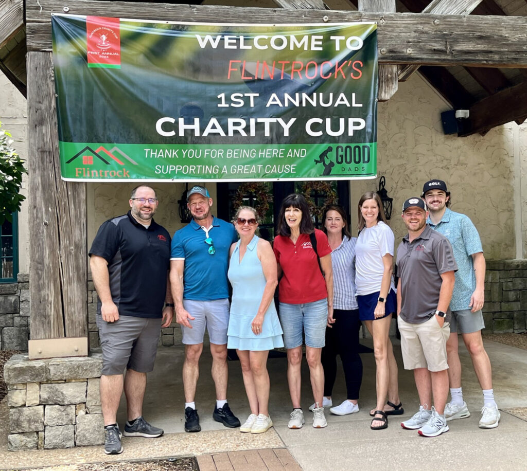 A group of happy golfers pose for a photo outdoors under a banner that says "welcome to Flintrock first annual Charity Cup"