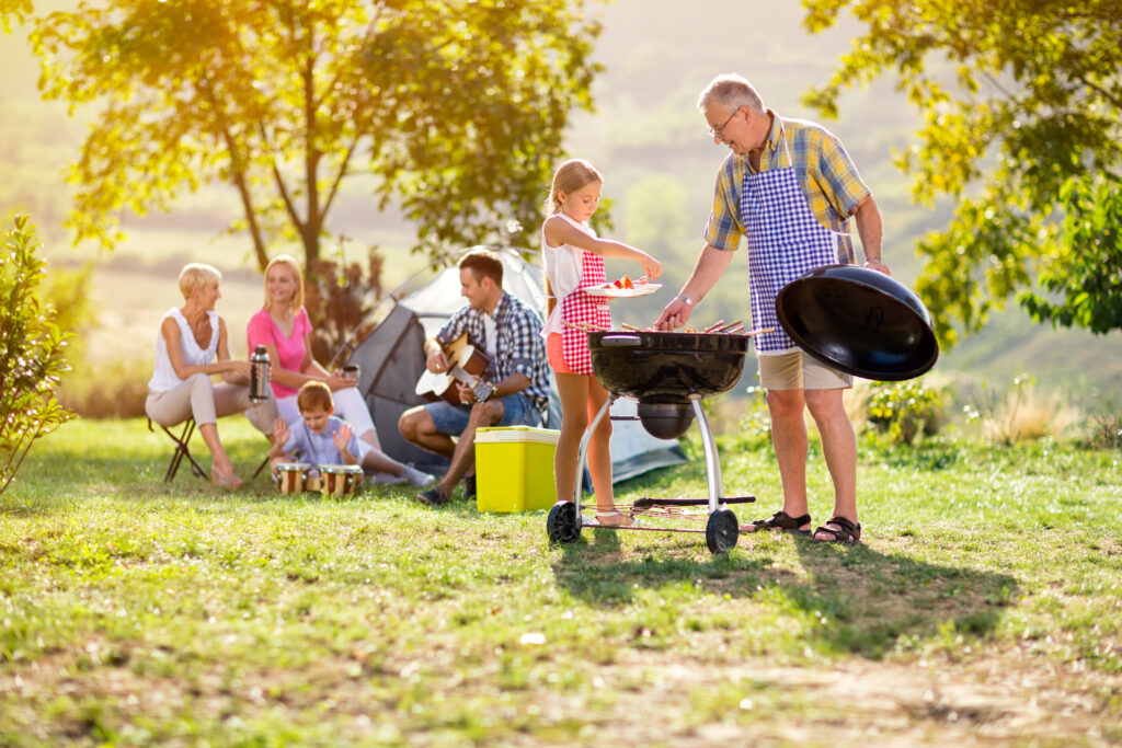 A happy, multi-generational family sits in a lush, green park while a grandfather and his granddaughter, both wearing aprons, barbeque foods at a grill.