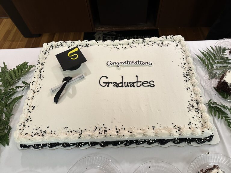 A cake with black and white icing decorated with a graduation cap and diploma. Words on the cake read "congratulations graduates"