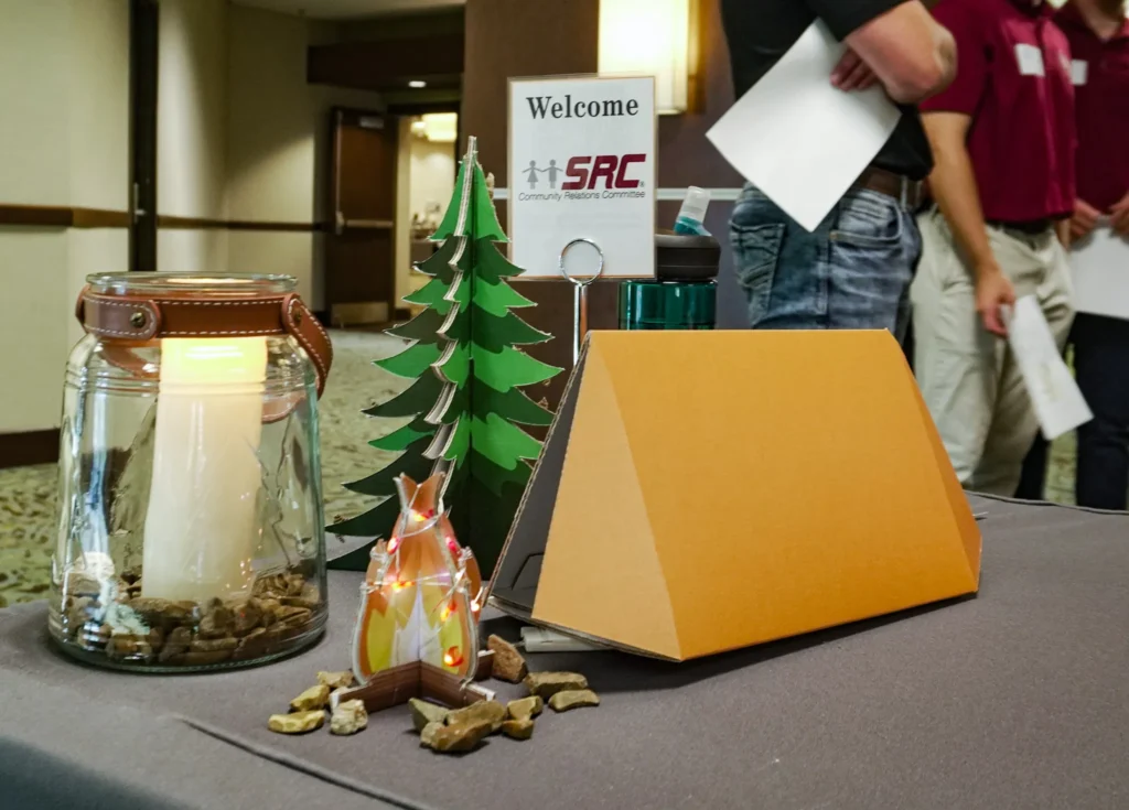 A close-up of table decorations at the check-in area in the lobby: a glass jar holding a lit candle and cardboard cut-outs of an orange camping tent, a small campfire and a pine tree surrounded by river rock.