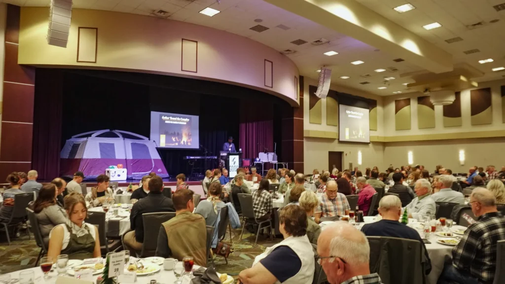 A wide-shot of a ballroom filled with people seated at tables: Radio personality J Fotsch stands at the podium and speaks into a microphone on stage.
