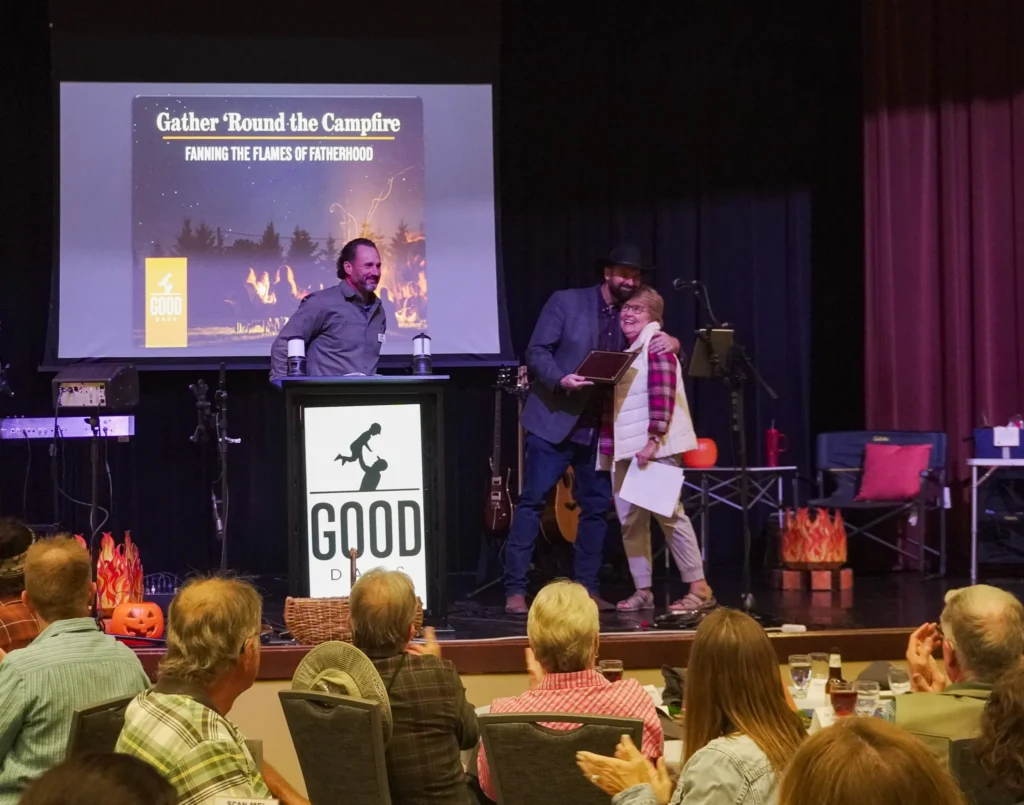 Good Dads founder and Director Dr. Jennifer L. Baker hugs emcee J Fotsch as she receives a plaque recognizing her for ten years of hard work at Good Dads; board president Joshua Wemple stands at the podium.