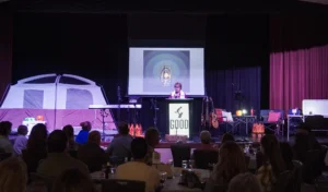 Dr. Jennifer L. Baker, Good Dads founder and director, stands at the podium on stage at the fundraising dinner, surrounded by camping decorations and an old-fashioned lantern flickering on the screen behind her.