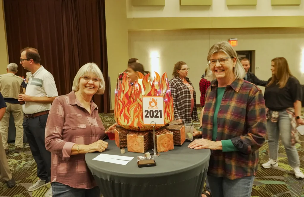 Good Dads volunteers Diane and Ingrid smile for a photo while standing at a trivia table decorated with a large cardboard campfire.