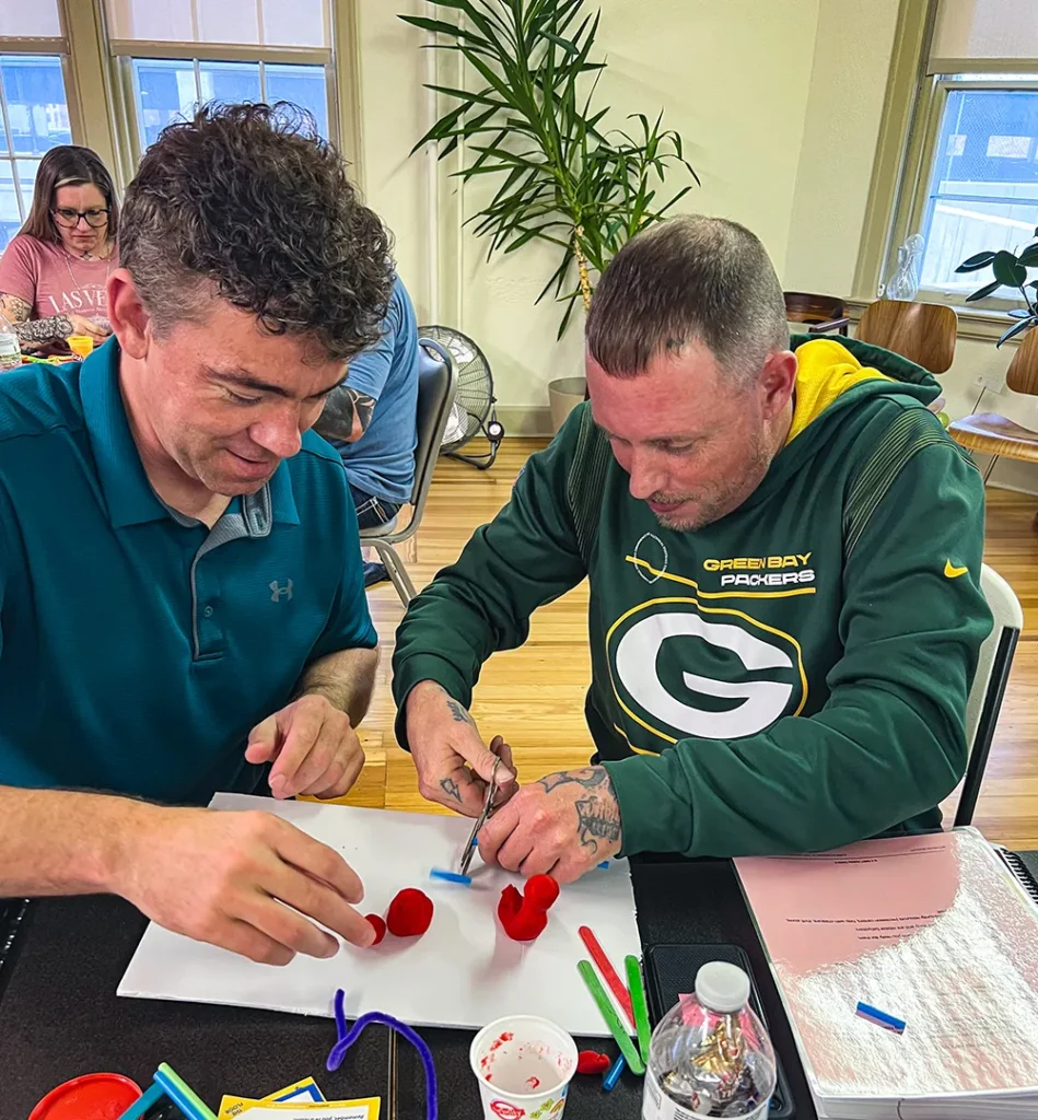 Two men, both attendees at Good Dads' training camp in November 2025, sit next to each other and collaborate to make small Play-Doh and pipe cleaner sculptures to represent a healthy relationship as part of the Within My Reach curriculum training.