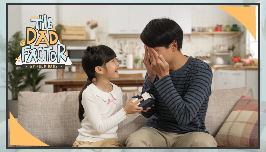 The Dad Factor: a father and daughter sit on a couch. The daughter holds out a present while the father covers his eyes.