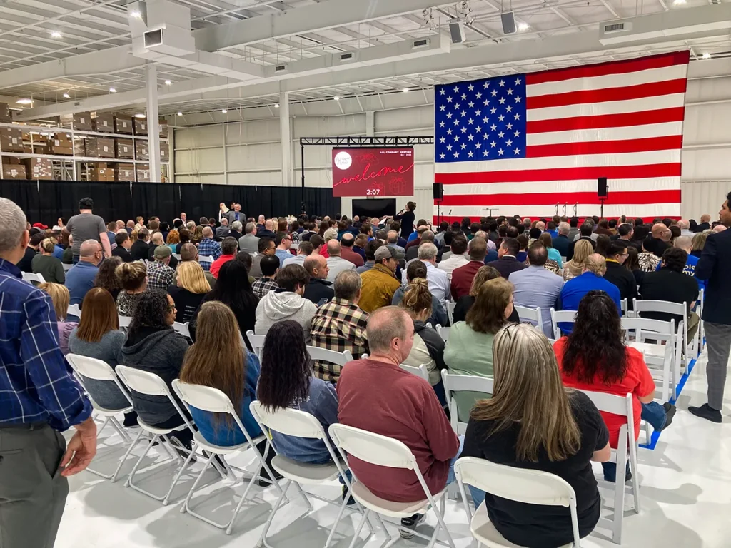 A large crowd of 350+ attendees seated at the DMP manufacturing headquarters faces a stage with a giant American flag backdrop, with Good Dads director Dr. Jennifer L. Baker on stage.