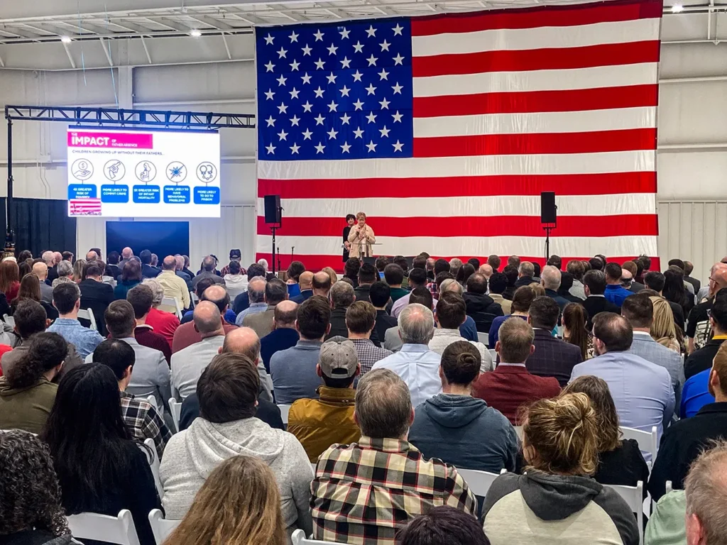 A large crowd of 350+ attendees seated at the DMP manufacturing headquarters faces a stage with a giant American flag backdrop, with Good Dads director Dr. Jennifer L. Baker on stage.