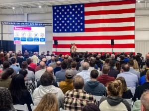 A large crowd of 350+ attendees seated at the DMP manufacturing headquarters faces a stage with a giant American flag backdrop, with Good Dads director Dr. Jennifer L. Baker on stage.