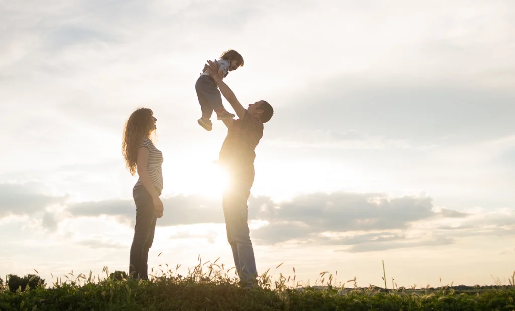 A family of three stand in a field, back-lit by the setting son. The dad holds his baby up in the air, while the mom stands nearby.