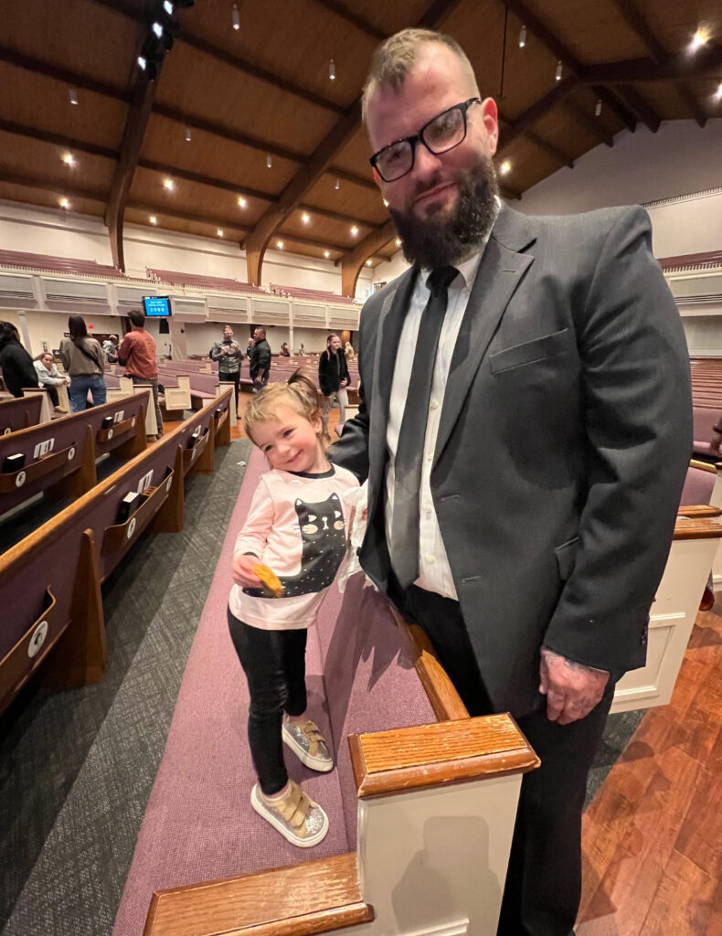 Ryan Warren wears a suit for a Victory Mission graduation ceremony, his 2-year-old daughter stands next to him.