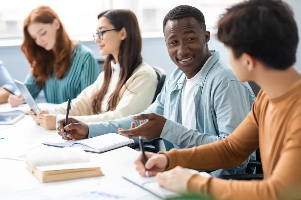A young Black man sits at a table with another man and two women, who all work together to learn in a classroom setting.