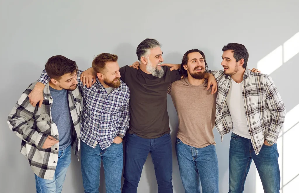 A group of five happy men, standing in a line next to each other, laughing joyously with their arms around each other's shoulders.