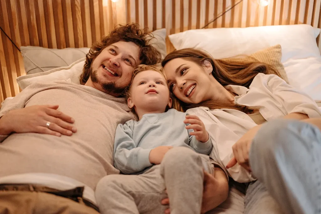 A happy young family, a dad, a mom and a toddler boy, lay in bed together to relax