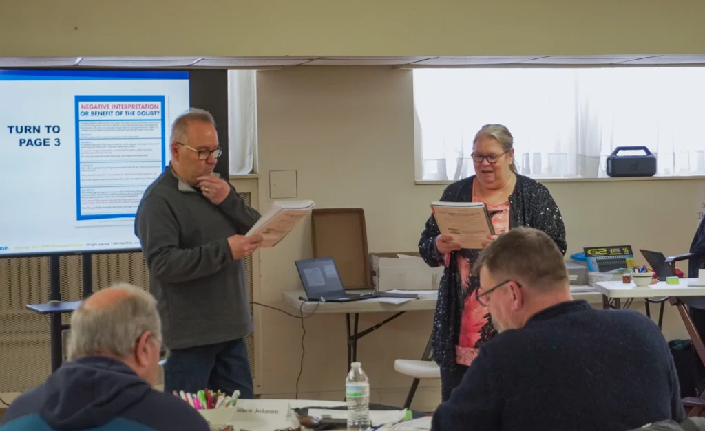 Facilitators Tim and Cindy, standing at the front of the room, practice reading aloud a scenario from their workbooks at the January 2026 Good Dads Plus Us Training Camp in Springfield MO.