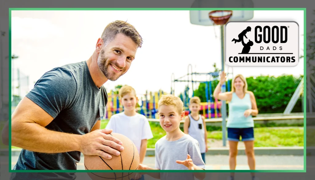 Good Dads Communicators: A fit-looking dad with a beard and salt-and-pepper hair holds a basketball and smiles mischievously, while his three sons and wife stand, out of focus, across the basketball court in a park.