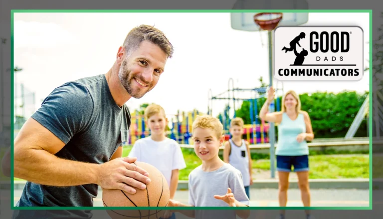 Good Dads Communicators: A fit-looking dad with a beard and salt-and-pepper hair holds a basketball and smiles mischievously, while his three sons and wife stand, out of focus, across the basketball court in a park.
