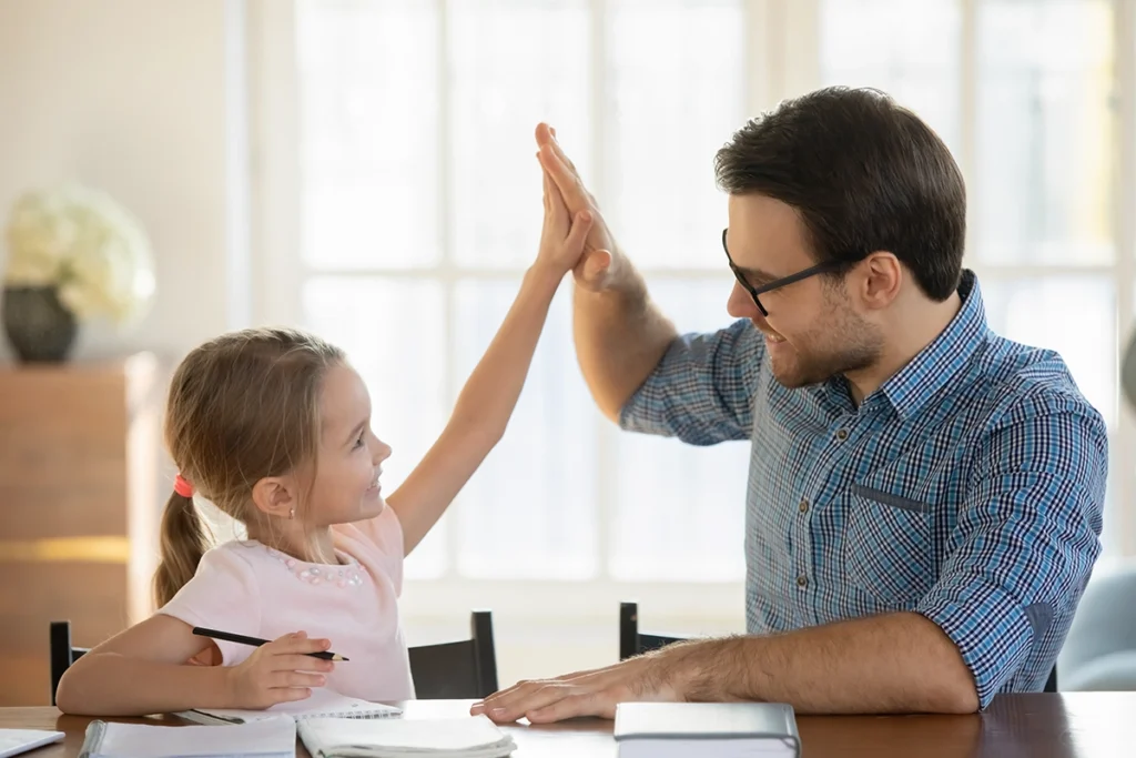 A dad and his young daughter, sitting indoors at a table in front of a stack of papers, possibly the girl's homework. They turn to each other, smiling, and high-five.