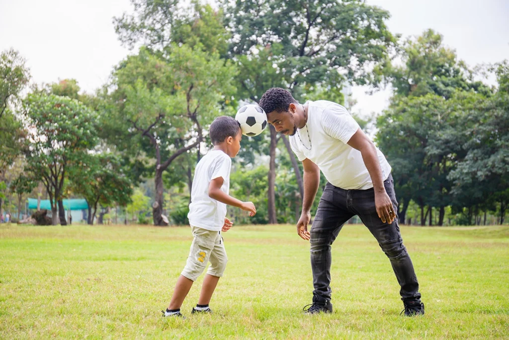 A dad wearing a white t-shirt and black jeans stands in a green field with his young son. Both hold a soccer ball between their heads.