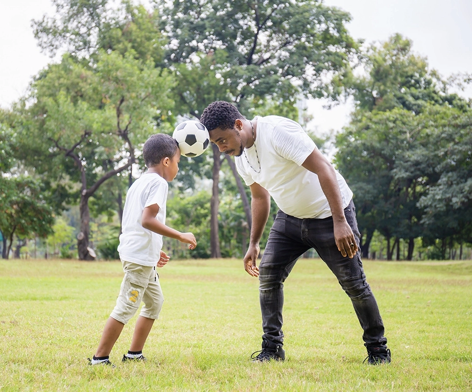 A dad wearing a white t-shirt and black jeans stands in a green field with his young son. Both hold a soccer ball between their heads.