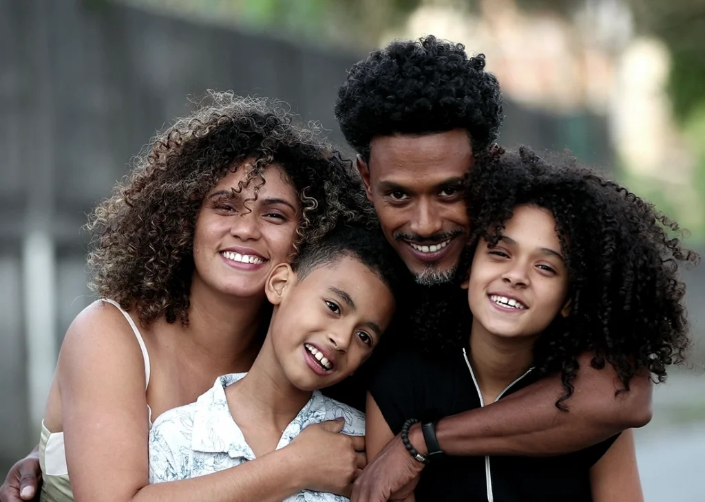 A happy, multi-racial family of four, including a dad, mom, son and daughter, stand close to one another for a group hug.