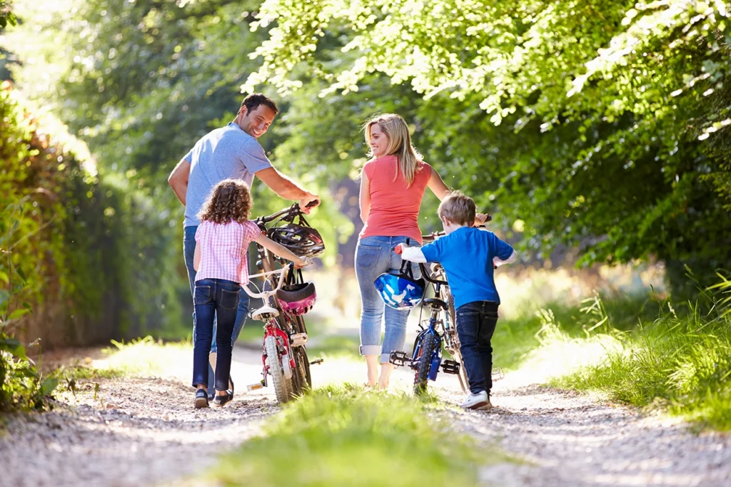 A family of four—a dad, mom, son and daughter—walk their bicycles along a gravel path surrounded by lush greenery.