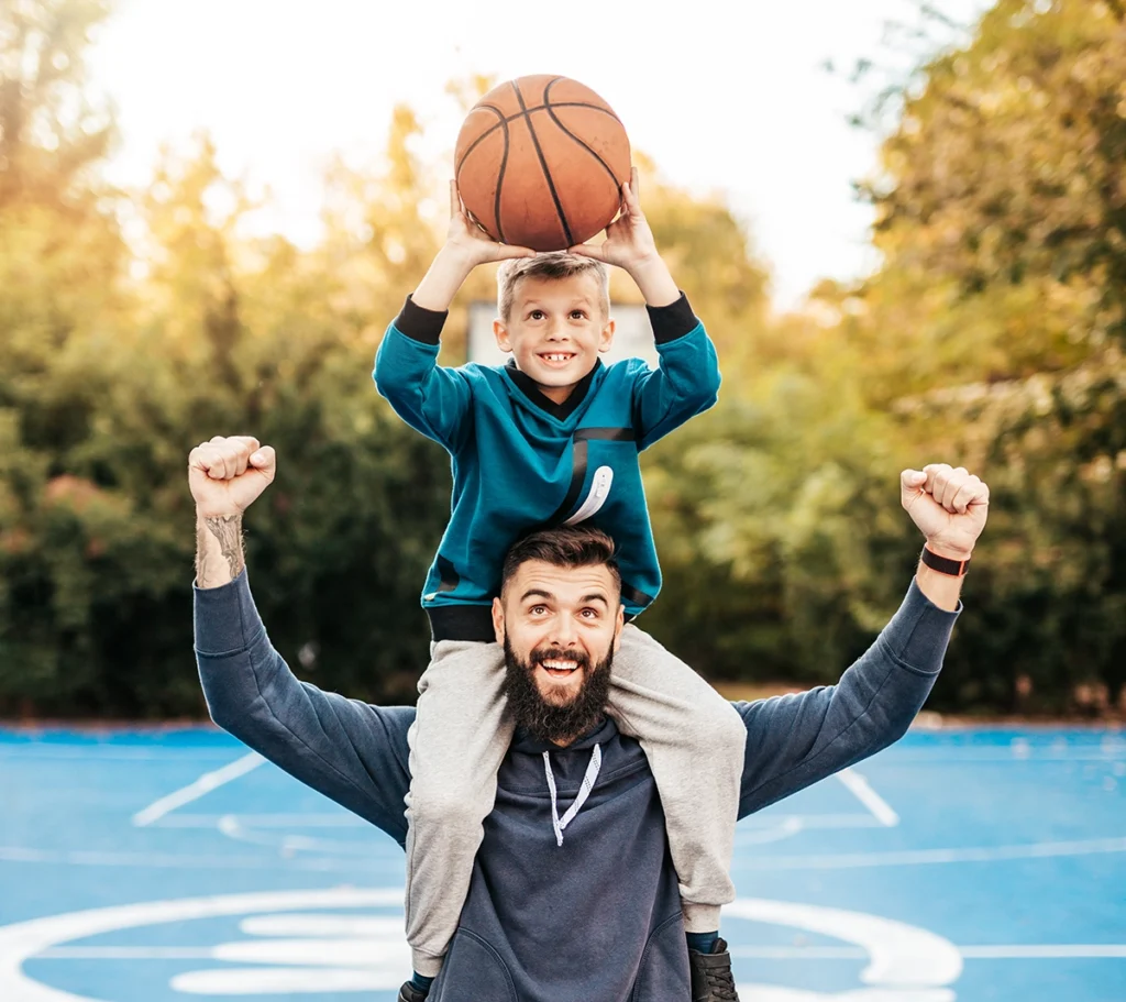 A happy dad, his arms in the air triumphantly, stands on a basketball court and holds his young son on his shoulders, while the boy excitedly hoists a basketball in the air.