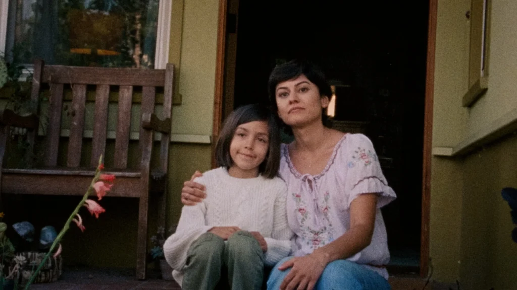 A mother and young daughter sit together on a porch, neutral expressions on their faces. The mother's arm is wrapped around the young daughter.