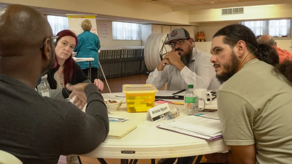 Good Dads facilitators, seated at round tables, intently collaborate and discuss a scenario during a Good Dads Training Camp activity in September 2026.