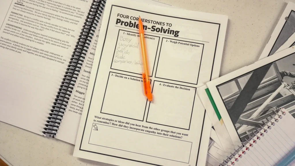 A close-up photo of a participant workbook at the September 2026 Good Dads Training Camp: It says "Four Cornerstones to Problem-Solving" and an orange mechanical pencil lays across the page.