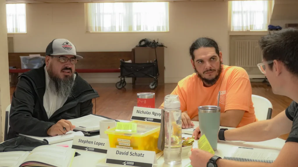 Good Dads facilitators, seated at round tables, intently collaborate and discuss a scenario during a Good Dads Training Camp activity in September 2026.